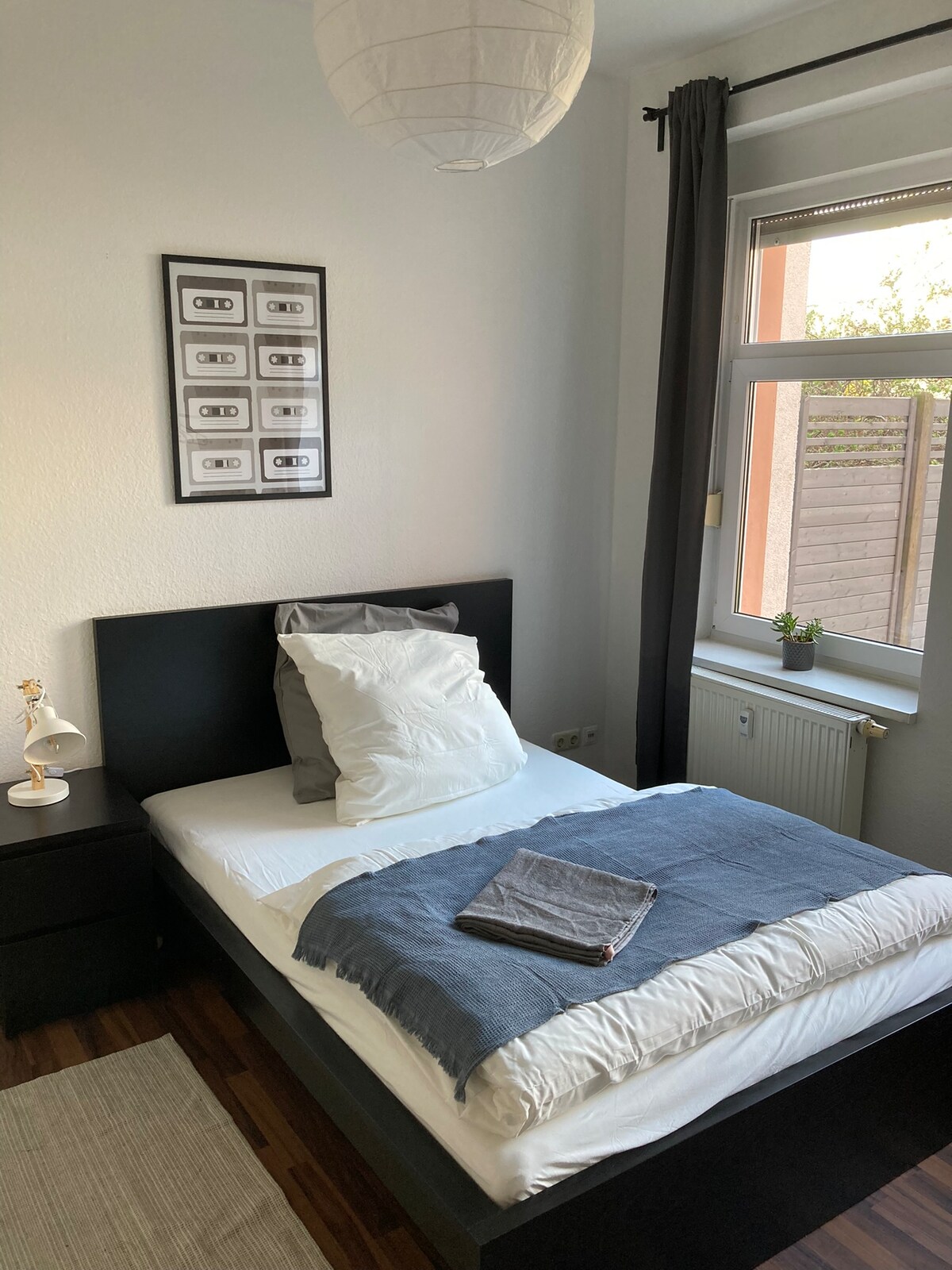 A cozy bedroom features a spacious bed adorned with white linens and a blue throw. A bedside table holds a small lamp, and framed artwork is displayed above the bed. Natural light enters through a large window, complemented by dark curtains.