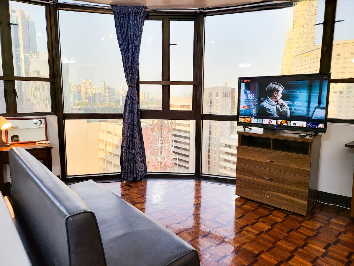 A spacious living area showcases large windows providing panoramic views of the city skyline. A comfortable sofa faces a flat-screen TV, positioned on a wooden media console. Natural light fills the space, with curtains elegantly framing the windows.