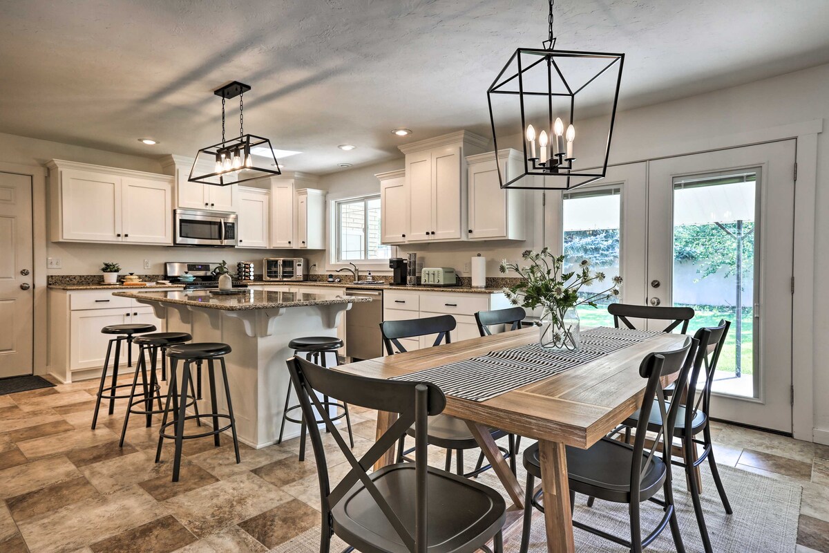 The modern kitchen showcases white cabinetry, stainless steel appliances, and a large island with bar seating. A dining table with cross-back chairs is positioned next to glass doors that lead to the backyard, allowing natural light to fill the space.
