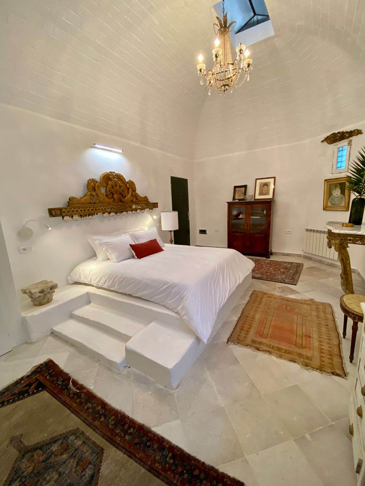 A spacious sleeping area features a plush bed with a white coverlet, elevated on steps. A chandelier hangs from the ceiling, providing soft lighting. Decorative elements include an ornate headboard and a vintage cabinet, complemented by various rugs on the stone floor.
