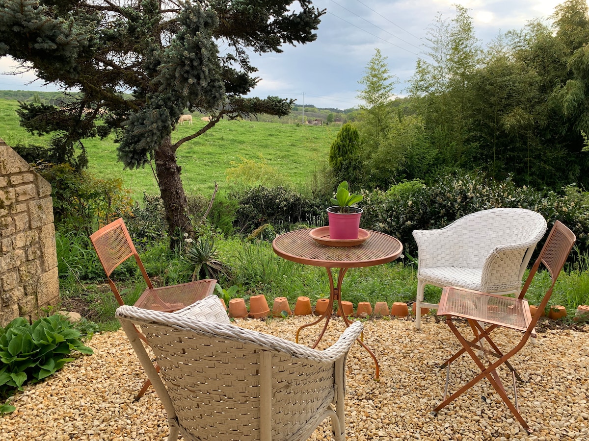 An outdoor seating area features two white chairs and a round table with a potted plant at its center. The stone path is surrounded by greenery and showcases distant rolling hills, providing a serene view of nature.