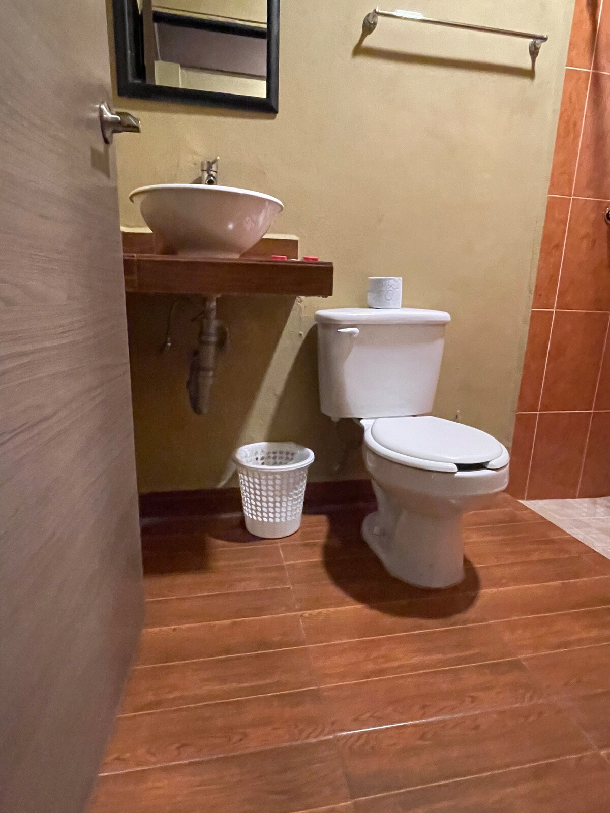 The bathroom features wooden flooring and terracotta tiled walls. A contemporary white toilet is positioned beside a small waste basket. A sleek, round sink is mounted on a wooden countertop, accompanied by a framed mirror above it. A shower area is also visible in the background.