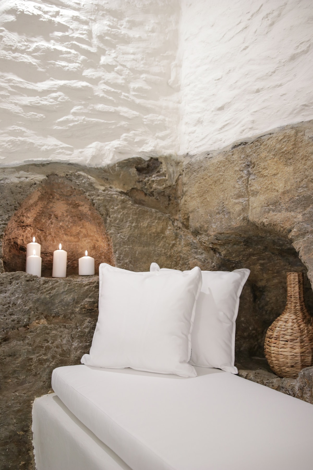 A serene corner features a cozy seating area with soft white cushions placed against a natural stone wall. Soft candlelight from three candles enhances the tranquil ambiance, while a woven basket adds a subtle decorative touch.