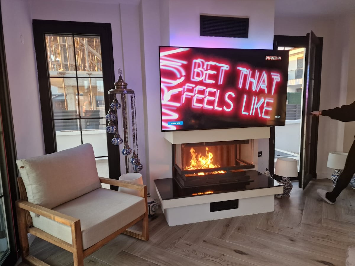 A contemporary living area features a modern fireplace beneath a large television displaying vibrant artwork. Natural light enters through expansive windows, enhancing the welcoming space. A comfortable chair is positioned nearby, contributing to a cozy atmosphere.