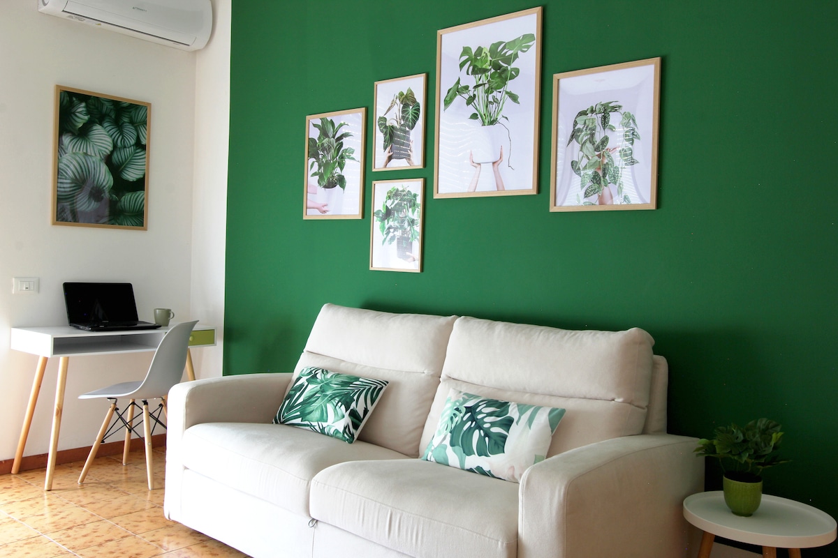 A spacious living area is highlighted by a light-colored sofa, adorned with decorative cushions featuring green patterns. A vibrant green wall serves as a backdrop for framed botanical artwork. A small desk with a chair is positioned in the corner, complementing the inviting space.