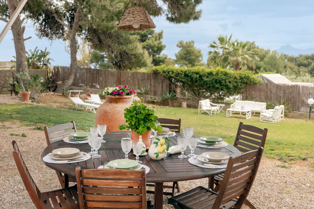 An outdoor dining area is set with a large wooden table, accompanied by eight matching chairs. Centered on the table is a terracotta pot filled with greenery. The spacious garden features lounge chairs in the background, framed by lush plants and trees.