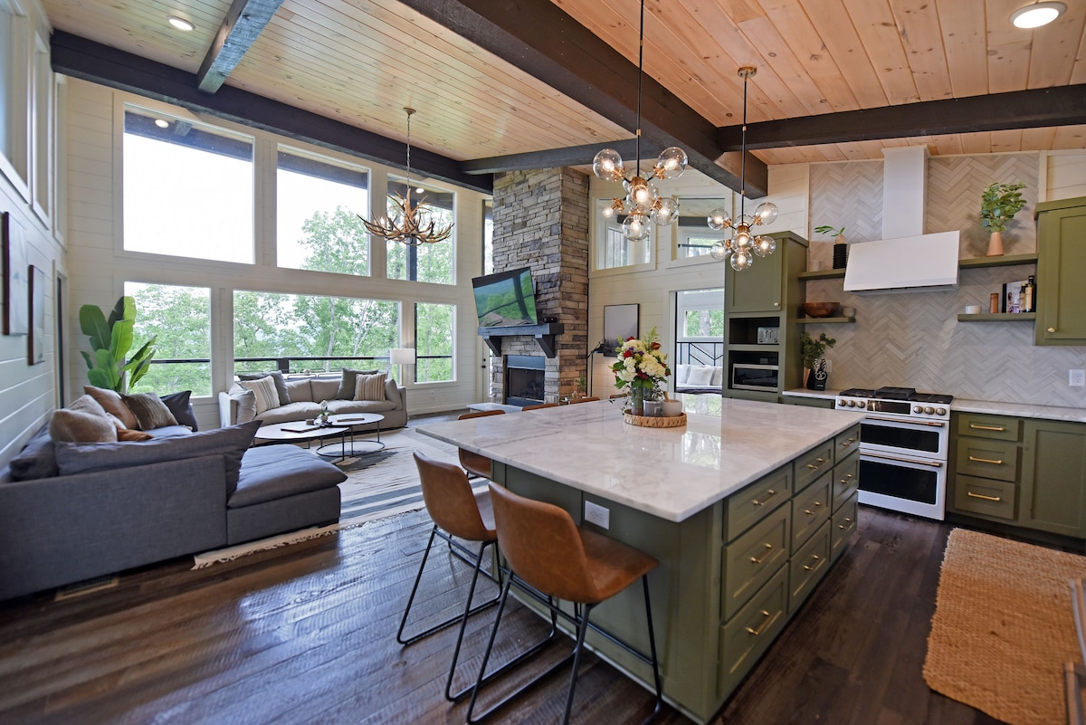 An open living space features an expansive kitchen island with modern bar stools. A comfortable seating area with soft sofas is situated near a stone fireplace and large windows, allowing natural light and scenic views of the outdoors.