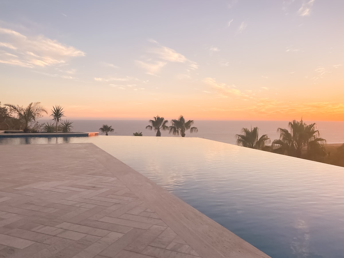 An infinity pool seamlessly merges with the horizon, reflecting the soft hues of a sunset. Lush palm trees frame the view, while the tranquil ocean is visible in the distance. The surrounding area features smooth stone tiles extending beyond the pool's edge.