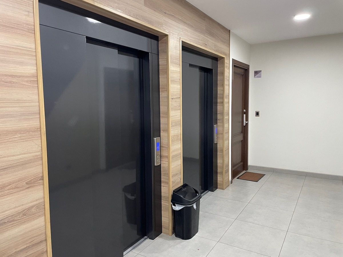 Two modern elevators are positioned side by side within a well-lit corridor, featuring wooden paneling on the walls. A trash bin is located nearby, and the floor is covered with neutral-colored tiles. A wooden door is visible, leading to an adjoining room.