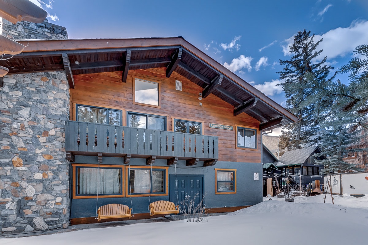 A two-story lodge featuring a charming wooden exterior with stone accents. The upper level has a balcony with two swings, set against a backdrop of snow-covered grounds and a clear blue sky. Surrounding trees complement the serene winter landscape.