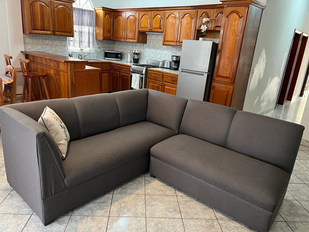 A sectional sofa with a simple design is positioned in the living area, accompanied by a throw pillow. The kitchen features wooden cabinets and modern appliances, including a refrigerator and stove, visible in the background. Natural light enters through a nearby window.