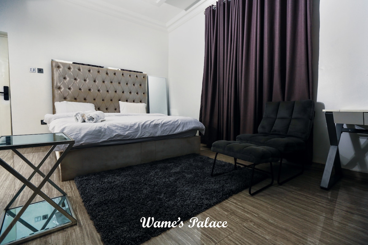 A spacious room showcases a large, tufted headboard bed, complemented by a soft area rug. A dark curtain provides privacy, while a modern chair adds seating. A glass table reflects light, creating an open and inviting environment for relaxation.