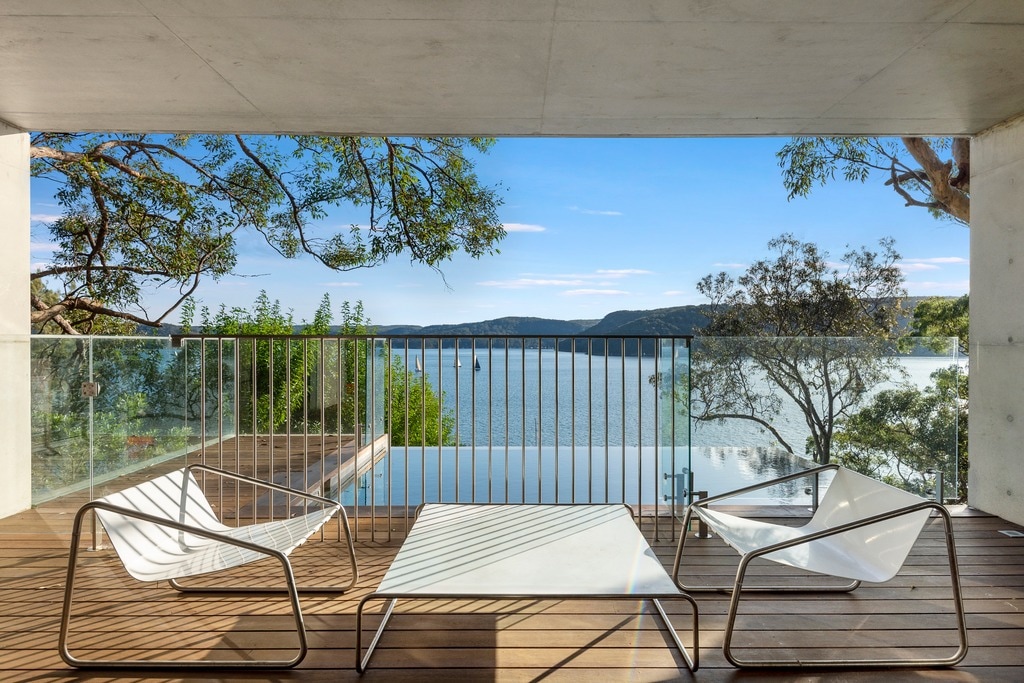 A spacious outdoor terrace features sleek white lounge chairs and a modern glass table. The view overlooks the tranquil waterfront, framed by green trees and distant hills. Natural light fills the area, enhancing the overall serene ambiance of the space.