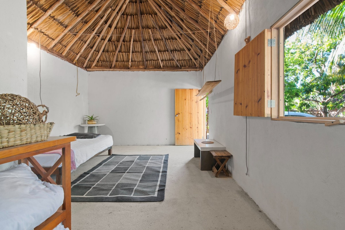 An airy room features a thatched ceiling and light-colored walls, creating a serene environment. Two single beds are positioned beside a window, allowing natural light to fill the space. A woven basket rests on a nearby table, and a small wooden stool is placed against the wall.