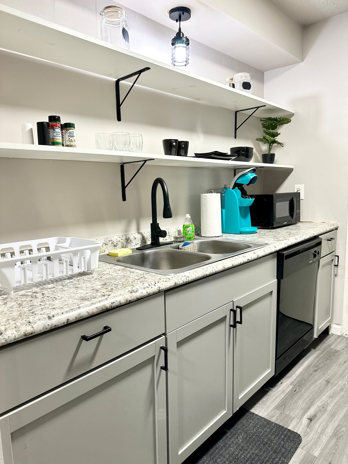 A modern kitchen area features light gray cabinetry with sleek black handles. A double sink is complemented by a black faucet, and ample counter space is provided by gray speckled countertops. Open shelves display various kitchen items, including a turquoise coffee machine and glassware.