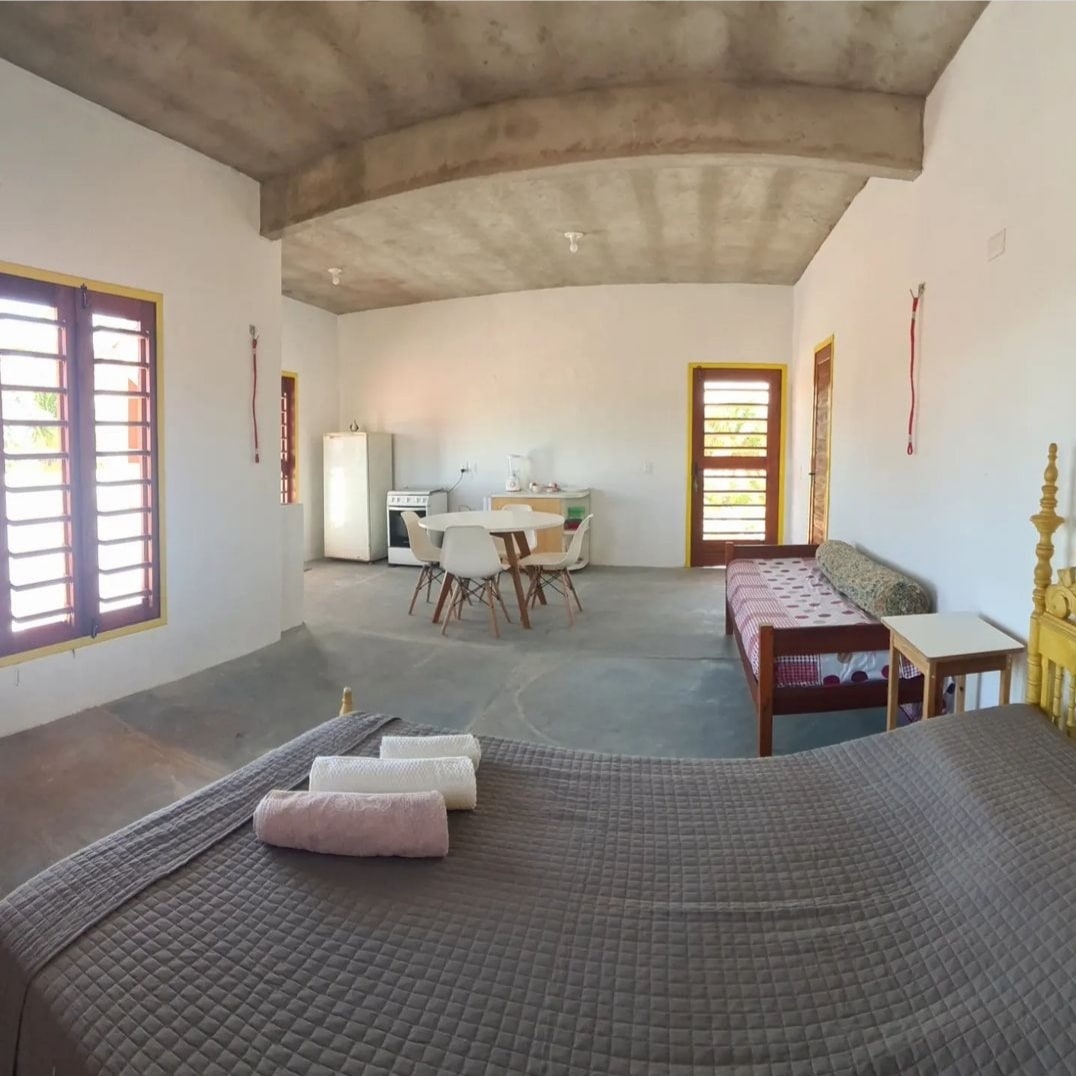 An open space with high ceilings features a spacious bed draped in a neutral quilt. A dining table for four and a cozy seating area are visible, while a kitchenette with a refrigerator is located in the background. Natural light filters through multiple windows.