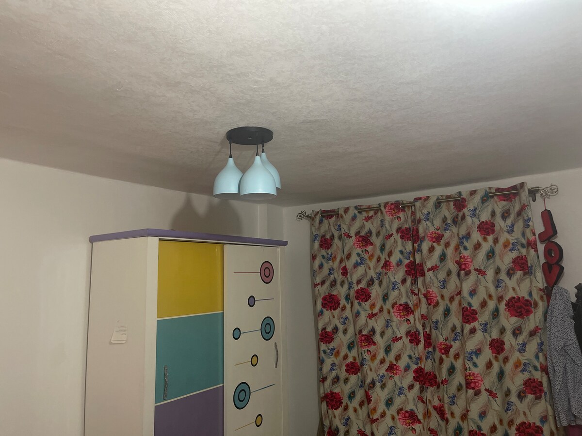 The room features a ceiling with a modern light fixture consisting of three blue shades. A colorful dresser with a geometric design stands against one wall. Floral-patterned curtains hang beside the dresser, providing a cheerful contrast to the neutral walls.