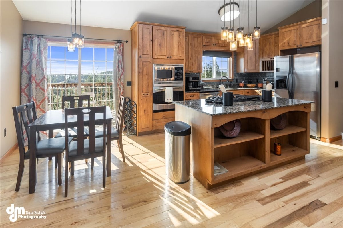 A modern kitchen area showcases warm wooden cabinetry and a spacious island with granite countertops. A dining table with four chairs is positioned beside a large window offering natural light. Stainless steel appliances are visible, along with a garbage can and open shelving featuring decorative items.