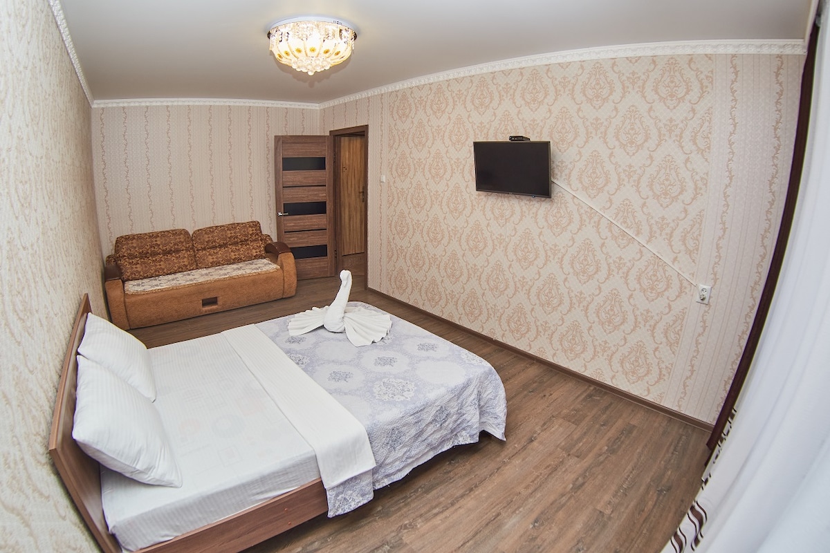 A spacious bedroom showcases a comfortable bed with neatly arranged pillows. A cozy sofa is positioned against a patterned wall, while a mounted television is visible opposite the bed. The room features warm wooden flooring under soft lighting from a ceiling fixture.