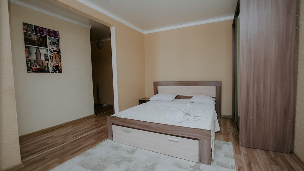 A minimalistic bedroom features a spacious bed with light linens and two pillows. A wooden headboard complements the room's neutral color scheme. A soft area rug covers part of the floor, and a large wardrobe is visible in the corner.