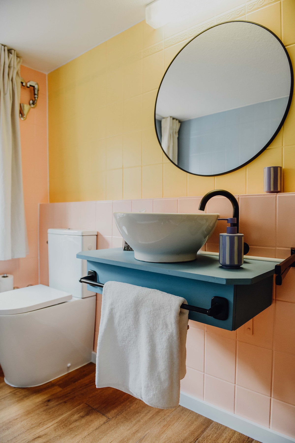 The bathroom features a modern toilet and a vessel sink mounted on a wall-mounted shelf. A round mirror above the sink reflects the vibrant yellow and pink tiled walls. A towel is neatly hung from the shelf, and a small candle adds a touch of decor.