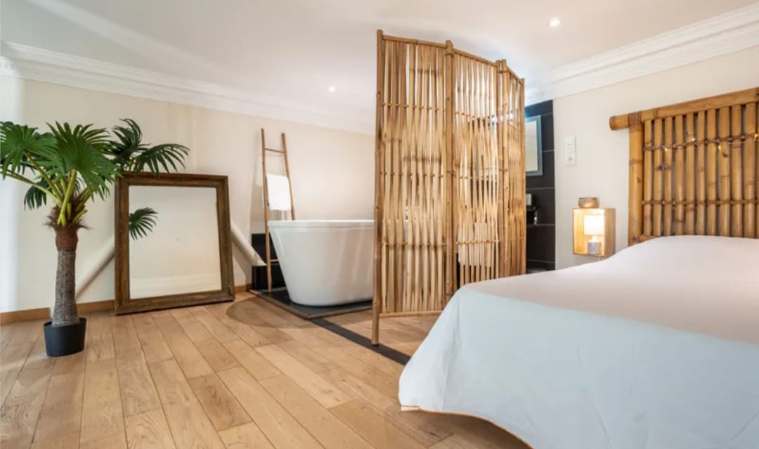 A serene space features a wooden bed with a light-colored duvet, complemented by a large mirror and a decorative plant. A bamboo partition separates an elegant bathtub from the sleeping area, enhancing the room's open layout and creating a peaceful ambiance.