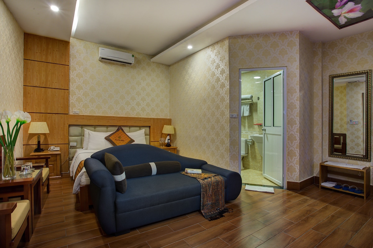A spacious hotel room features a comfortable sofa and a king-sized bed, decorated with neutral-coloured walls and wooden accents. The en-suite bathroom is visible through a glass door. Two lamps provide soft lighting, contributing to a relaxed ambiance.