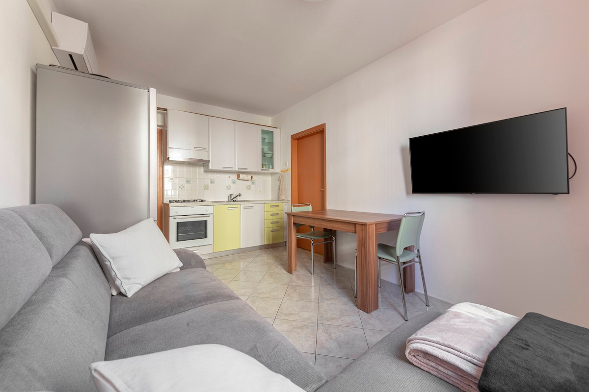 A modern living area features a gray sofa with cushions and a wooden dining table. A kitchenette with yellow accents is visible, and a flat-screen television is mounted on the wall. Natural light illuminates the space through the doorway.