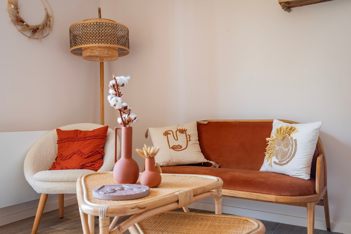 A cozy seating area features a rounded chair and a curved sofa adorned with decorative cushions. A woven lamp stands next to the sofa, illuminating the soft, neutral tones. A wooden coffee table displays a decorative plate and a simple floral arrangement.
