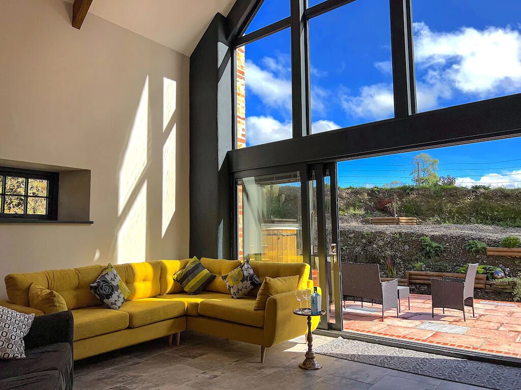 A spacious living area is illuminated by large floor-to-ceiling windows, showcasing the expansive outdoor space. A modern yellow sectional sofa provides ample seating, while sunlight casts dynamic shadows on the walls. The sliding glass doors open to a terrace with outdoor furniture.