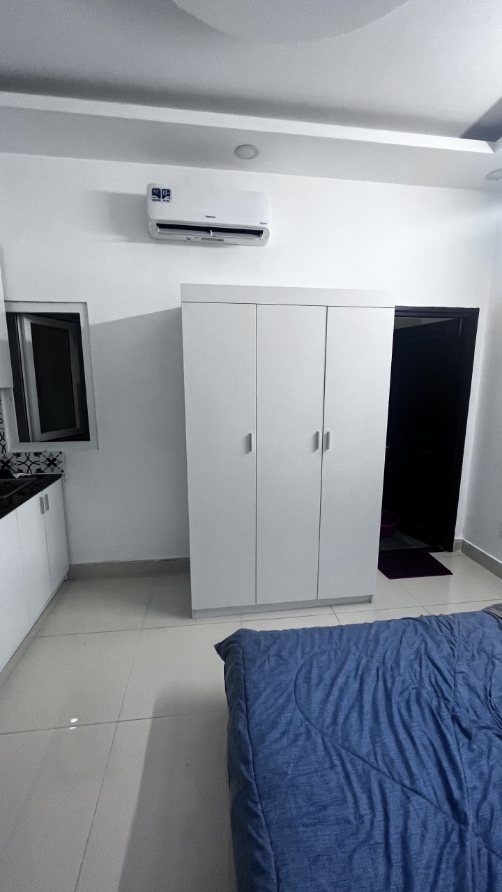 A compact room is presented with a spacious bed dressed in a blue coverlet. A white wardrobe stands against the wall, while an air conditioning unit is mounted above. A kitchenette with subtle cabinetry is visible alongside a window, providing natural light.