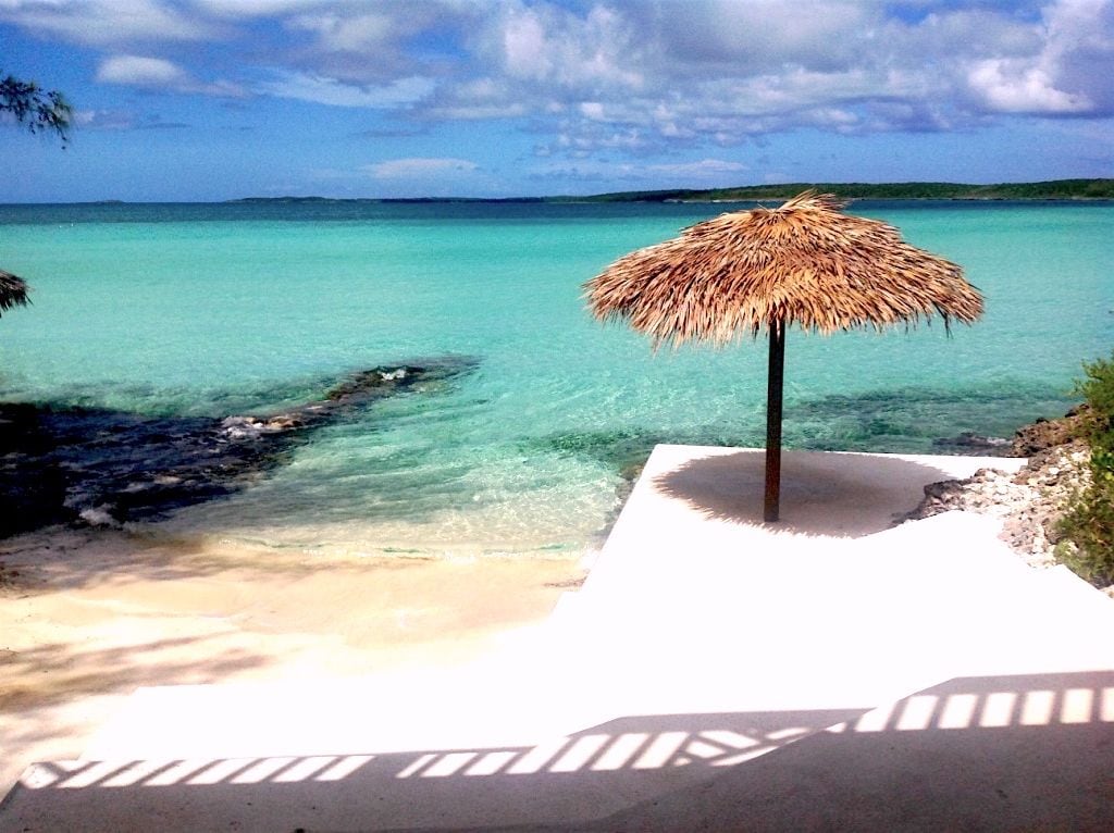 A thatched umbrella provides shade on a small private beach area, overlooking calm, turquoise waters. Gentle waves lap against a sandy shoreline, framed by lush greenery and smooth rocks. The clear sky reflects light onto the vibrant water, enhancing the serene coastal view.