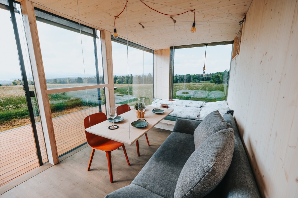 A wooden interior features large glass windows that provide views of the surrounding landscape. A cozy seating area is complemented by a dining table set for two. The space is filled with natural light, enhancing the tranquil atmosphere.