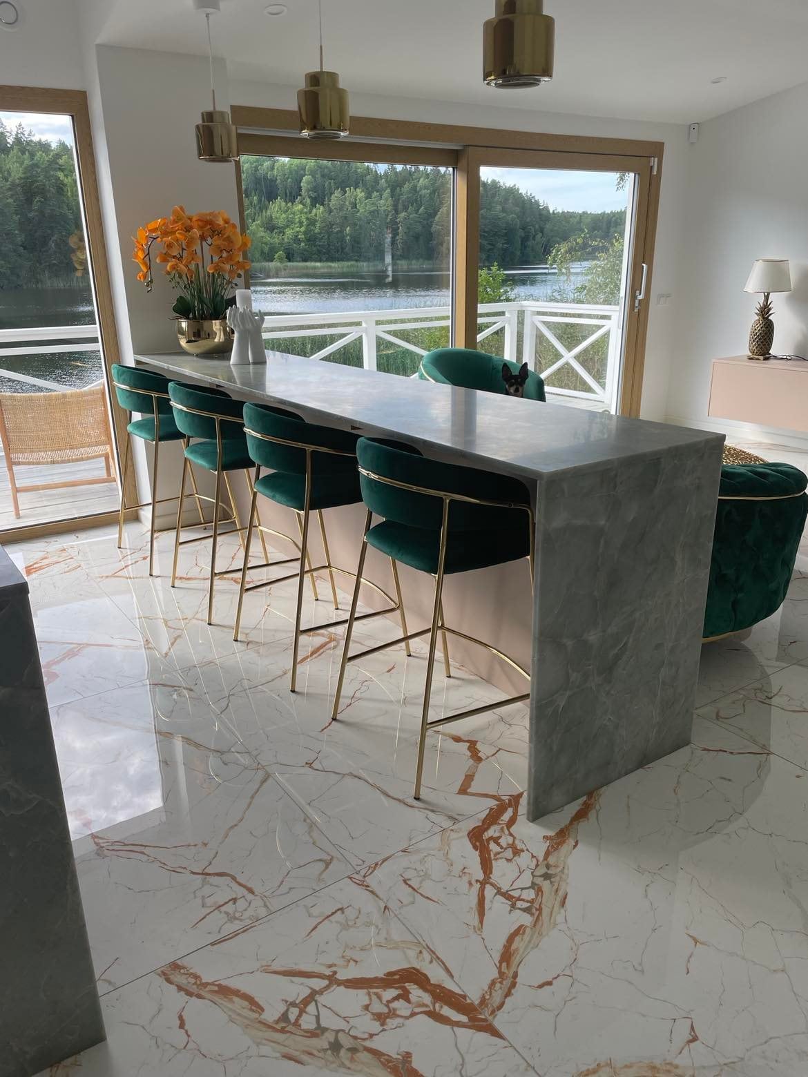 A modern dining area features a sleek marble countertop with four high-backed, green velvet stools. Large windows provide a view of the serene lake and surrounding trees, while warm lighting from stylish pendant lamps enhances the inviting atmosphere of the space.
