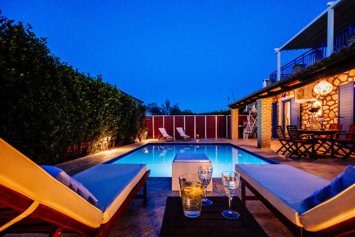 A private pool area is surrounded by lush greenery, softly illuminated by warm lighting. Comfortable lounge chairs are positioned beside the sparkling water, with a stylish table featuring glasses nearby. The stone house stands in the background under a serene twilight sky.
