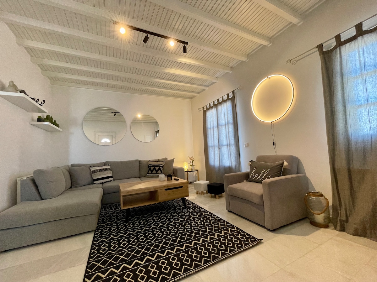 A bright living area features a sectional gray sofa, accompanied by a comfortable armchair. A wooden coffee table is centered on a patterned rug. Mirrors reflect natural light, while sheer curtains filter it slightly. An illuminated circular wall fixture adds subtle ambiance.