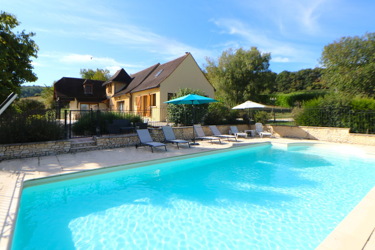 A private swimming pool is bordered by a sun terrace featuring multiple loungers and umbrellas. The house is visible in the background, surrounded by greenery. The tranquil blue water of the pool reflects the clear sky, offering a serene outdoor space.