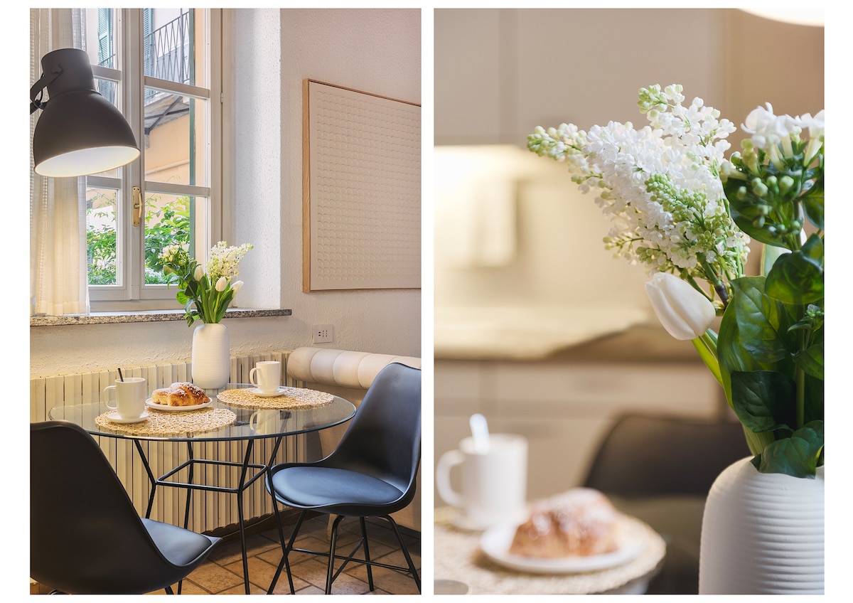 A dining area features a small round table set for two, adorned with plates, cups, and a pastry. A vase holds fresh flowers, adding a touch of greenery. The space is illuminated by a modern pendant light, with a window in the background offering a glimpse of the outdoors.