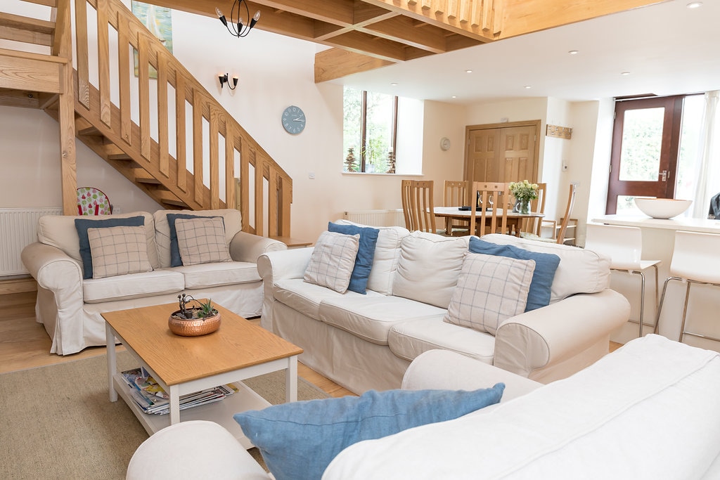 A spacious living area features two white sofas paired with blue accent pillows. A wooden coffee table sits in the center, surrounded by soft furnishings and natural light from large windows. A wooden staircase leads to the upper level, enhancing the open-plan design.