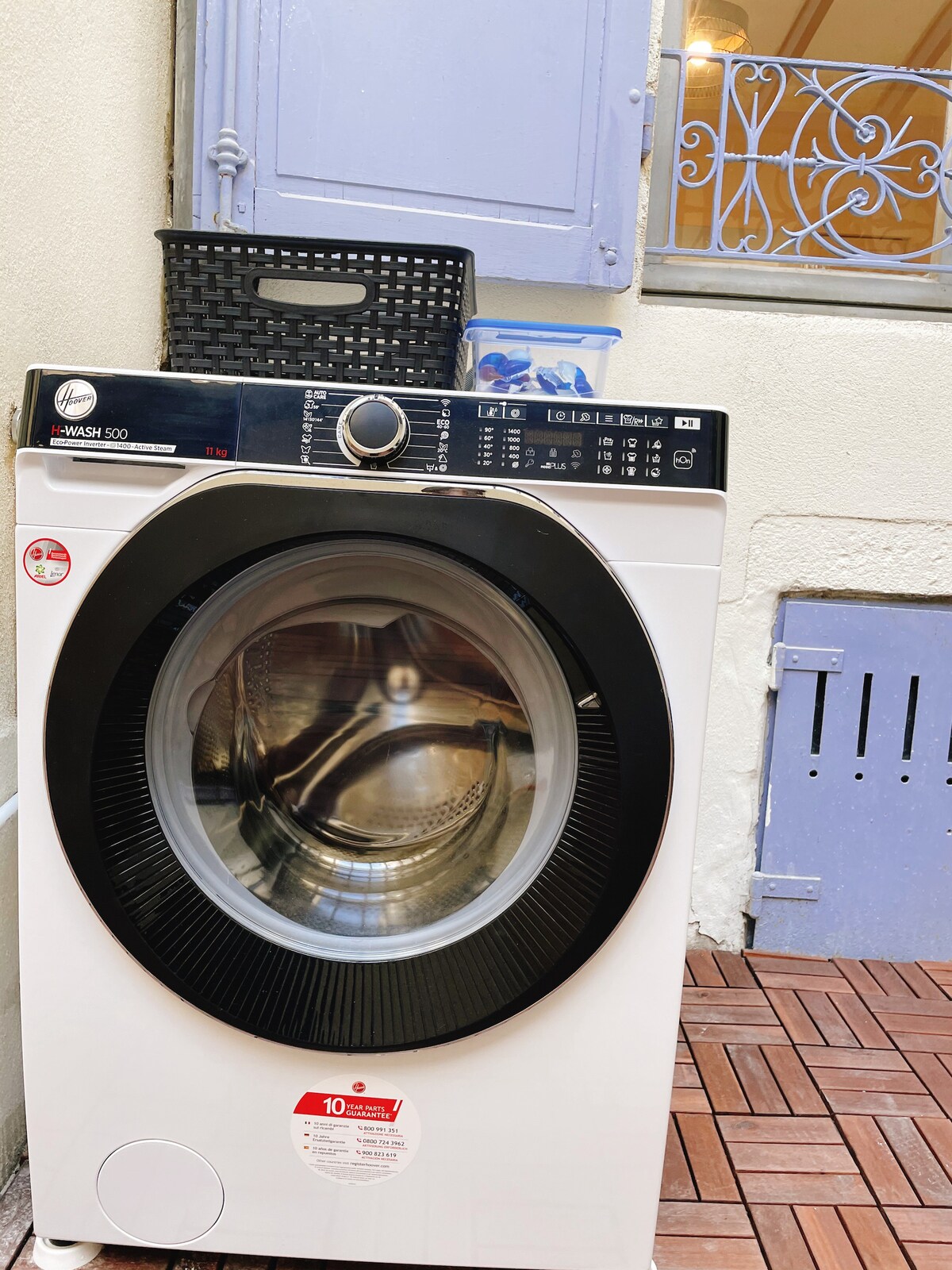 A washing machine is positioned on a tiled surface in a bright space. The glass door of the machine reveals the drum inside. Nearby, a woven basket and a partially open window with decorative metalwork is visible, contributing to the functional setting.