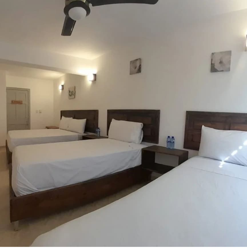 A bright guestroom showcases three spacious beds with light bedding arranged against a neutral wall. Simple wooden nightstands accompany each bed. Soft lighting is provided by wall-mounted fixtures, and two bottles of water are placed on one of the nightstands.