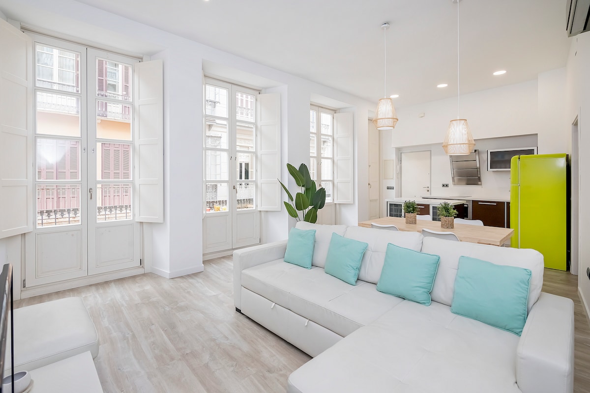 A spacious living area is highlighted by a light-colored sectional sofa adorned with turquoise cushions. Large windows allow natural light to fill the room, while open shutters provide a view of the outside. A modern kitchen with sleek appliances can be seen in the background.