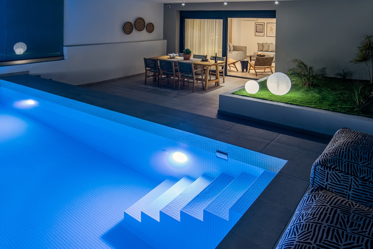 A serene evening view of a private swimming pool, highlighted by soft blue lighting. Adjacent, a dining area with a large table is visible, facing an interior space defined by gentle lighting and minimal decor. Potted plants and modern lighting accents enhance the outdoor ambiance.