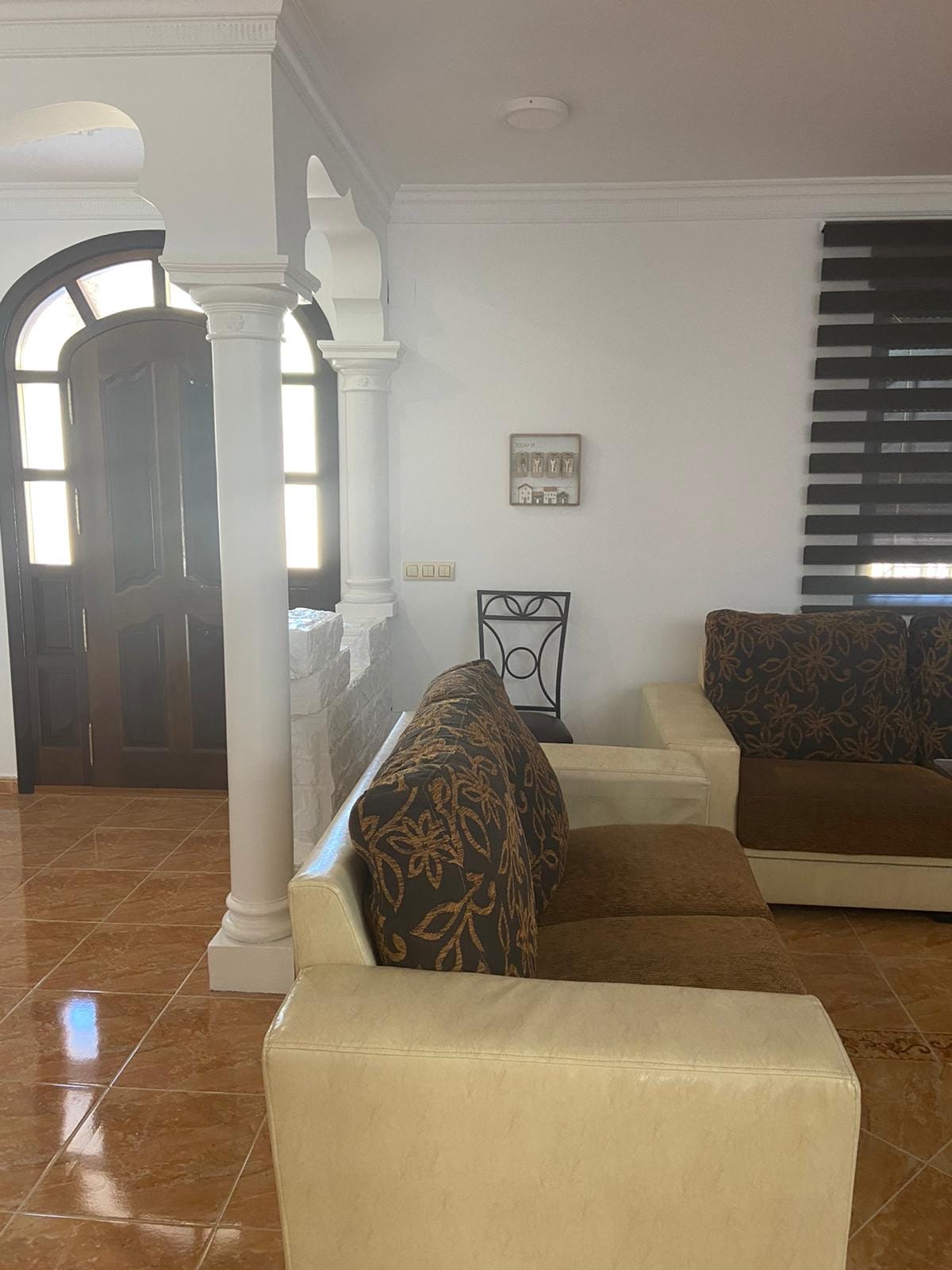 A living area features two sofas with contrasting fabrics, positioned adjacent to a decorative wall. Detailed columns frame an entrance leading outside. A black chair is positioned nearby, while light filters through window coverings, creating a softly lit environment.