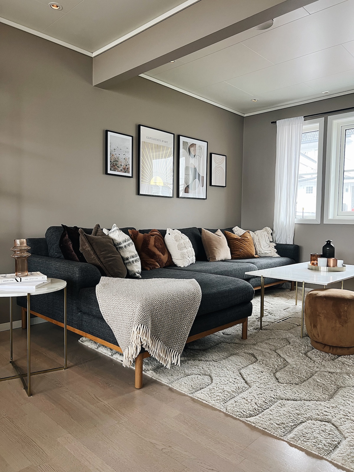 A modern living area features a dark sectional sofa adorned with a mix of decorative pillows in neutral tones. A light-colored coffee table is placed in front, complemented by soft textured rugs. Natural light enters through two windows, enhancing the cozy atmosphere.