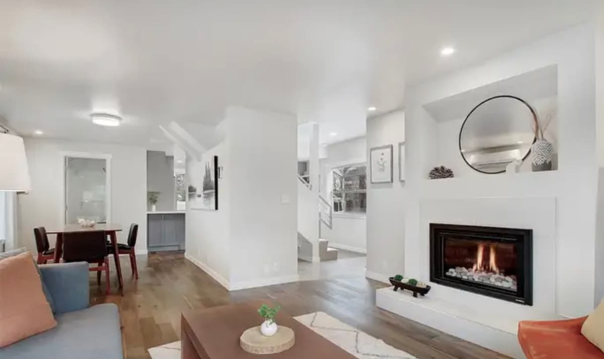 A modern living area features a comfortable sofa and armchair, with a coffee table positioned centrally. A fireplace is set into a white feature wall, reflecting soft light. Natural wood flooring extends throughout, while a dining area with a table and chairs is visible in the background.