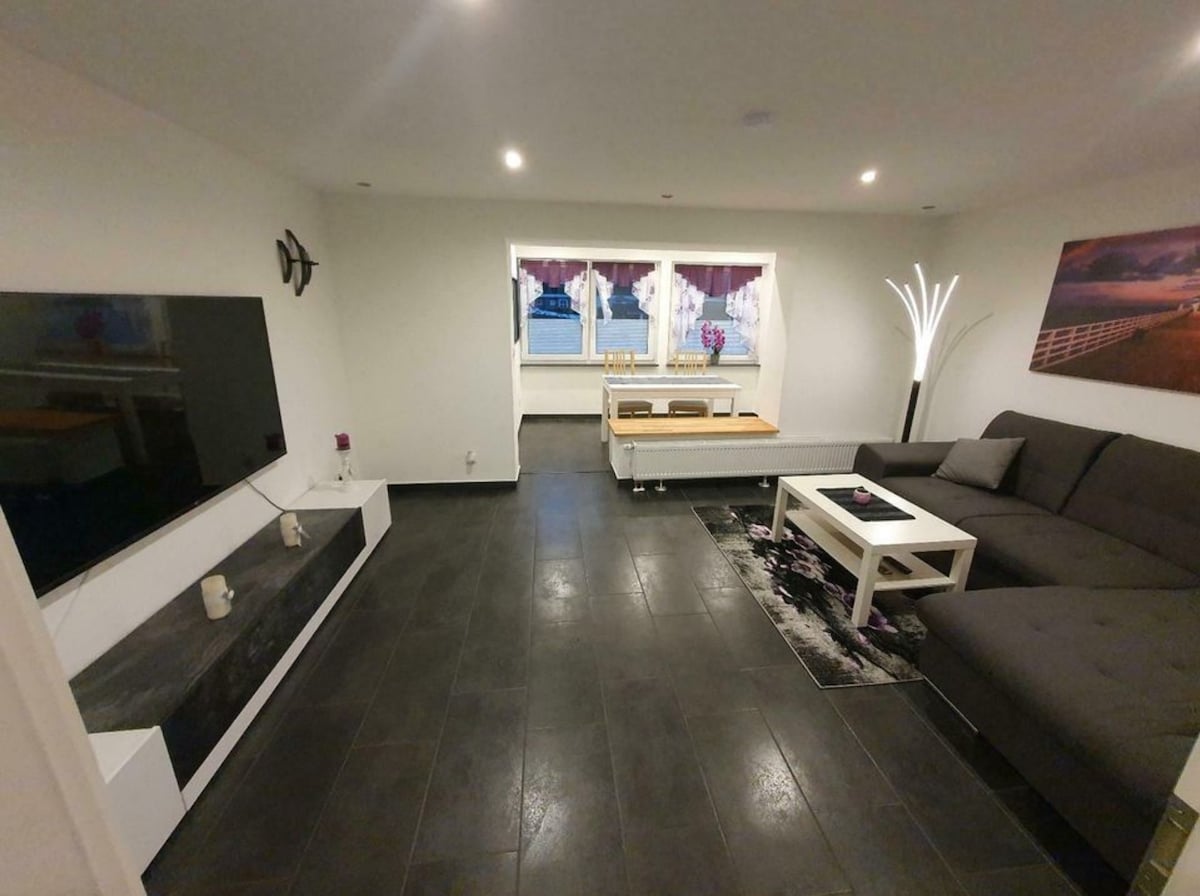 A modern living area is shown with a large sofa and a sleek coffee table at the center. A flat-screen television is mounted on the wall opposite. Natural light is filtered through large windows adorned with purple curtains, while a decorative floor lamp adds visual interest.