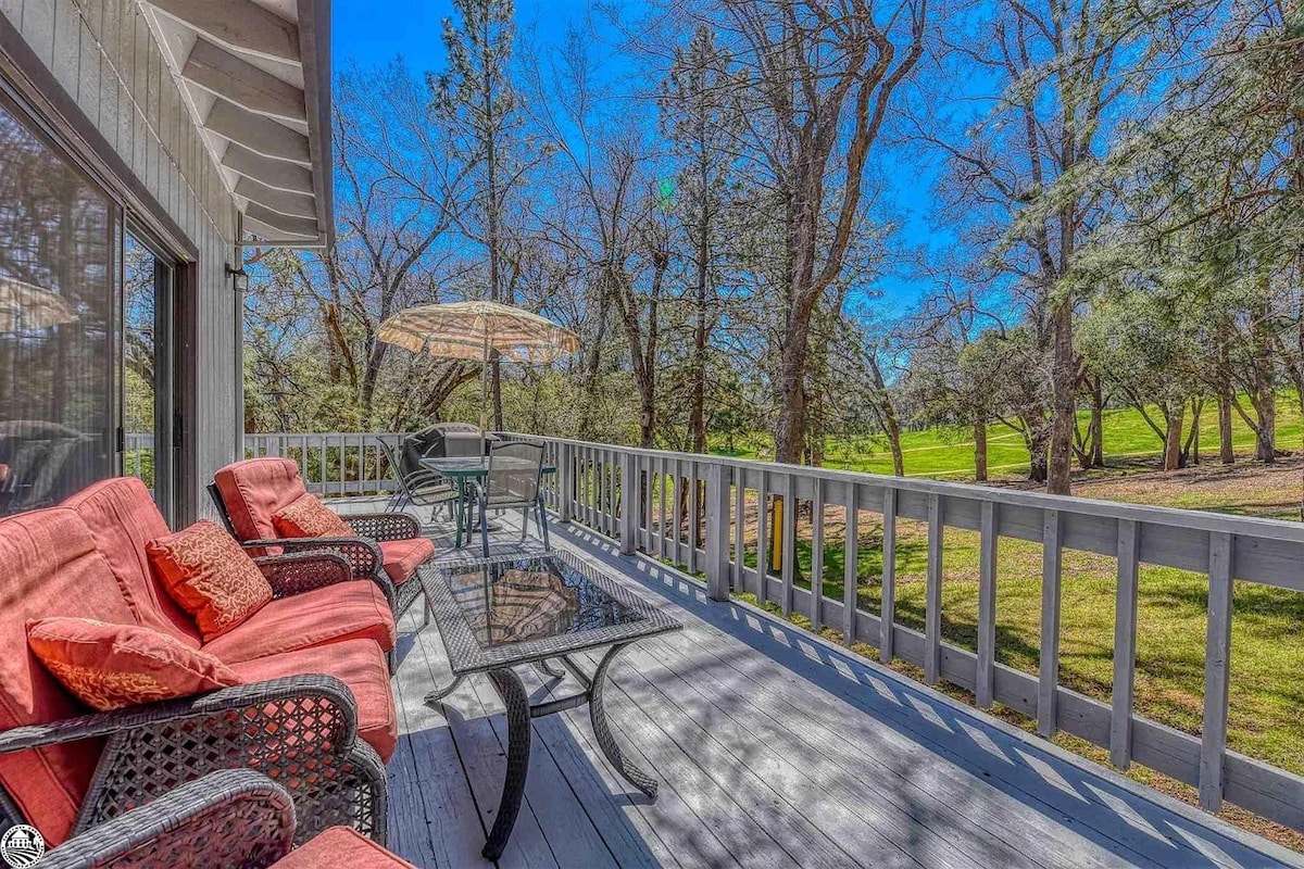 A spacious outdoor deck features cushioned seating arrangements and a glass-topped table. An umbrella provides shade for a dining set positioned nearby. The deck overlooks lush greenery and a golf course, framed by tall trees under a clear blue sky.