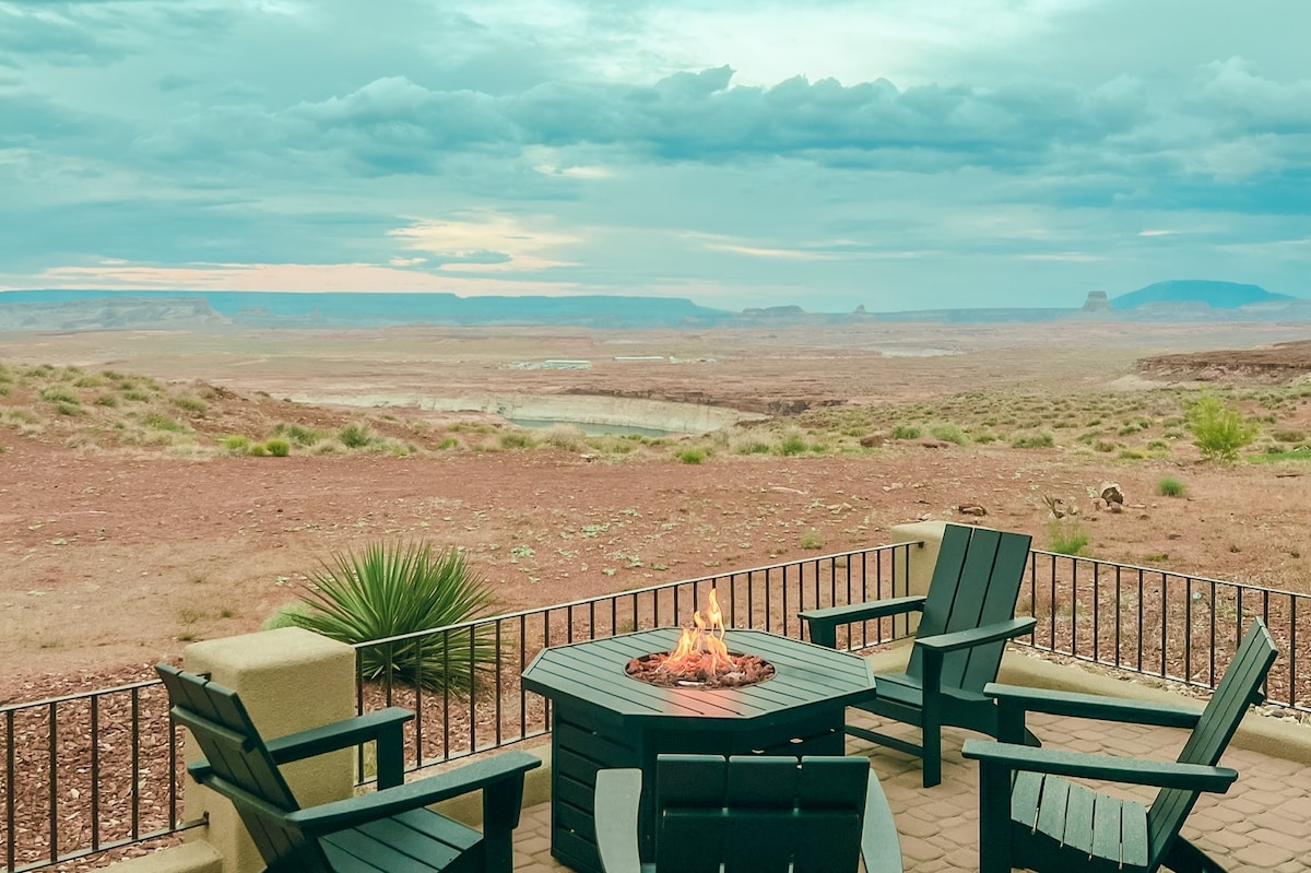 An outdoor patio features a Polywood dining table surrounded by four Adirondack chairs. A propane firepit sits at the center, offering warmth against the backdrop of expansive desert vistas and distant hills under a cloudy sky.