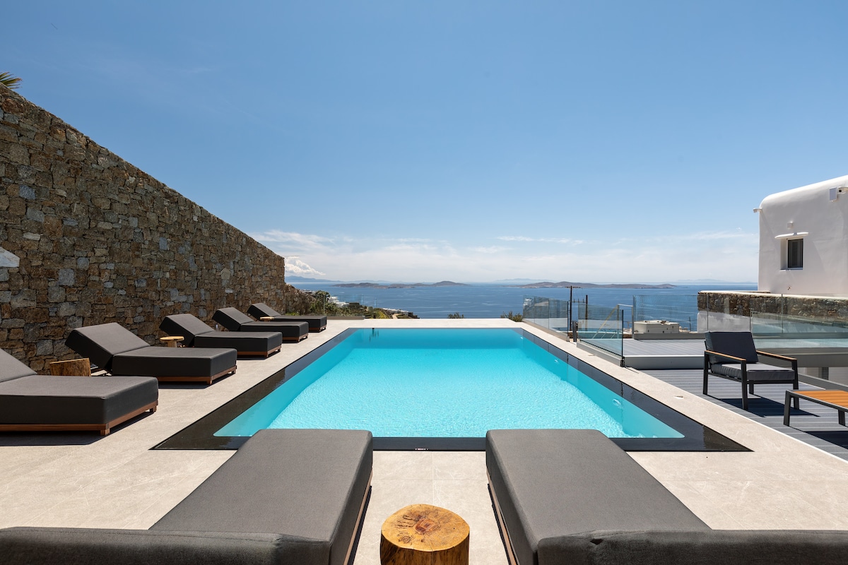 A sparkling blue pool is surrounded by sleek sun loungers, inviting relaxation. A stone wall lines one side, while expansive views of the sea and the horizon are visible in the distance under a clear sky.